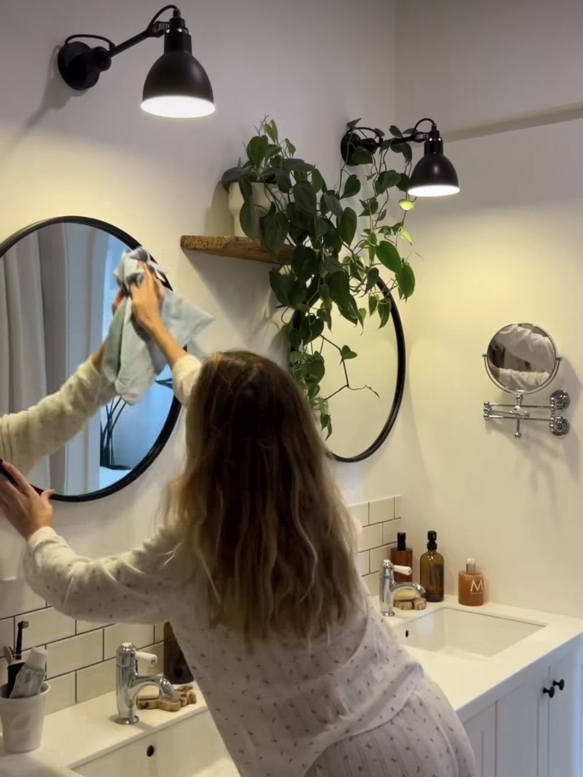 A person wiping a round bathroom mirror with a blue cloth in a bright room.