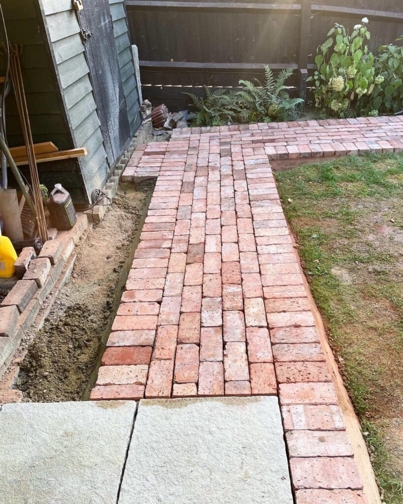 Red brick garden pathway laid in a herringbone pattern next to a sand trench.