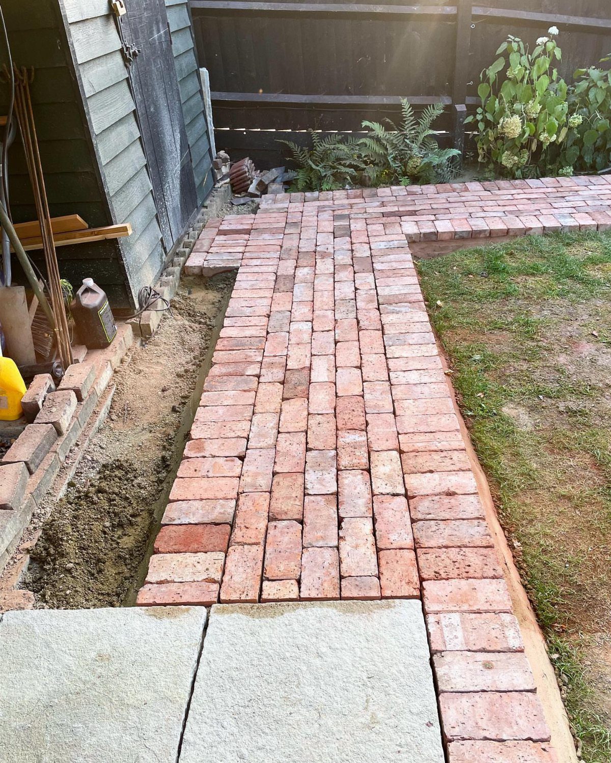 Red brick garden pathway laid in a herringbone pattern next to a sand trench.