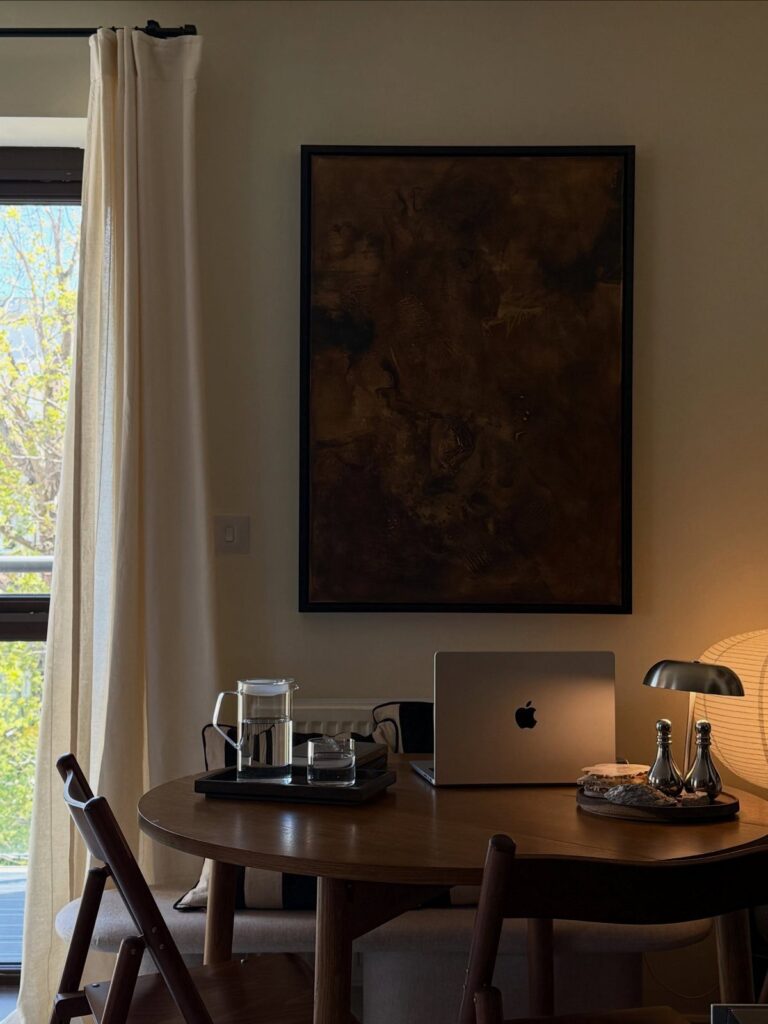 A wooden dining table with a laptop and lamp set against a wall with large abstract art.