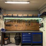 A wooden workbench with a grey top, blue tool chests, and various hand tools hanging on a wall.
