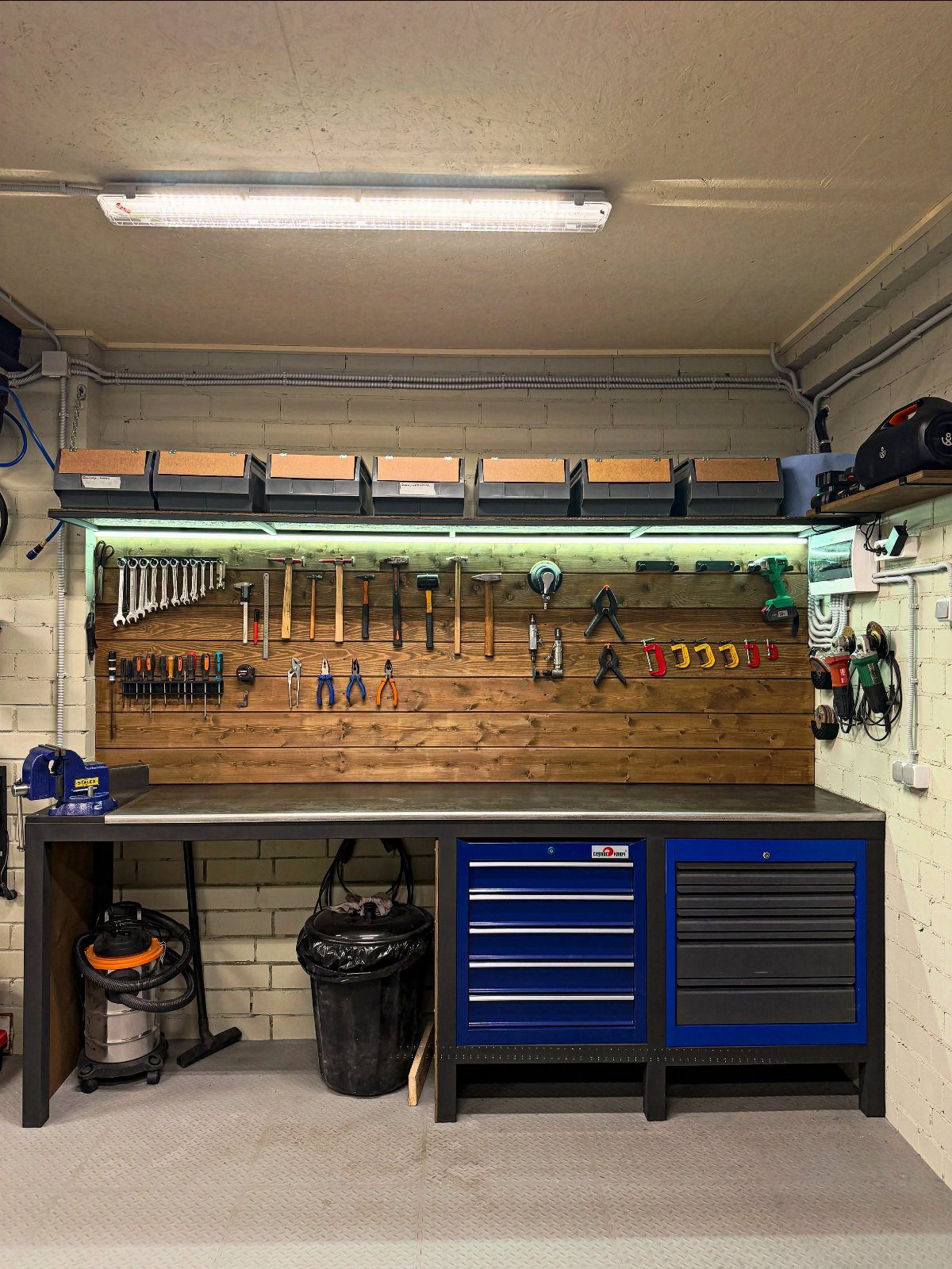 A wooden workbench with a grey top, blue tool chests, and various hand tools hanging on a wall.