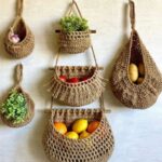 Woven jute hanging baskets containing garlic, plants, tomatoes, and citrus fruits mounted on a wall.