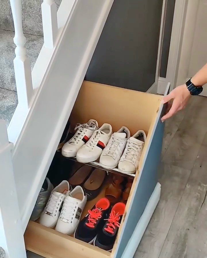 A wooden pull-out drawer filled with various pairs of shoes being pulled from under white stairs.